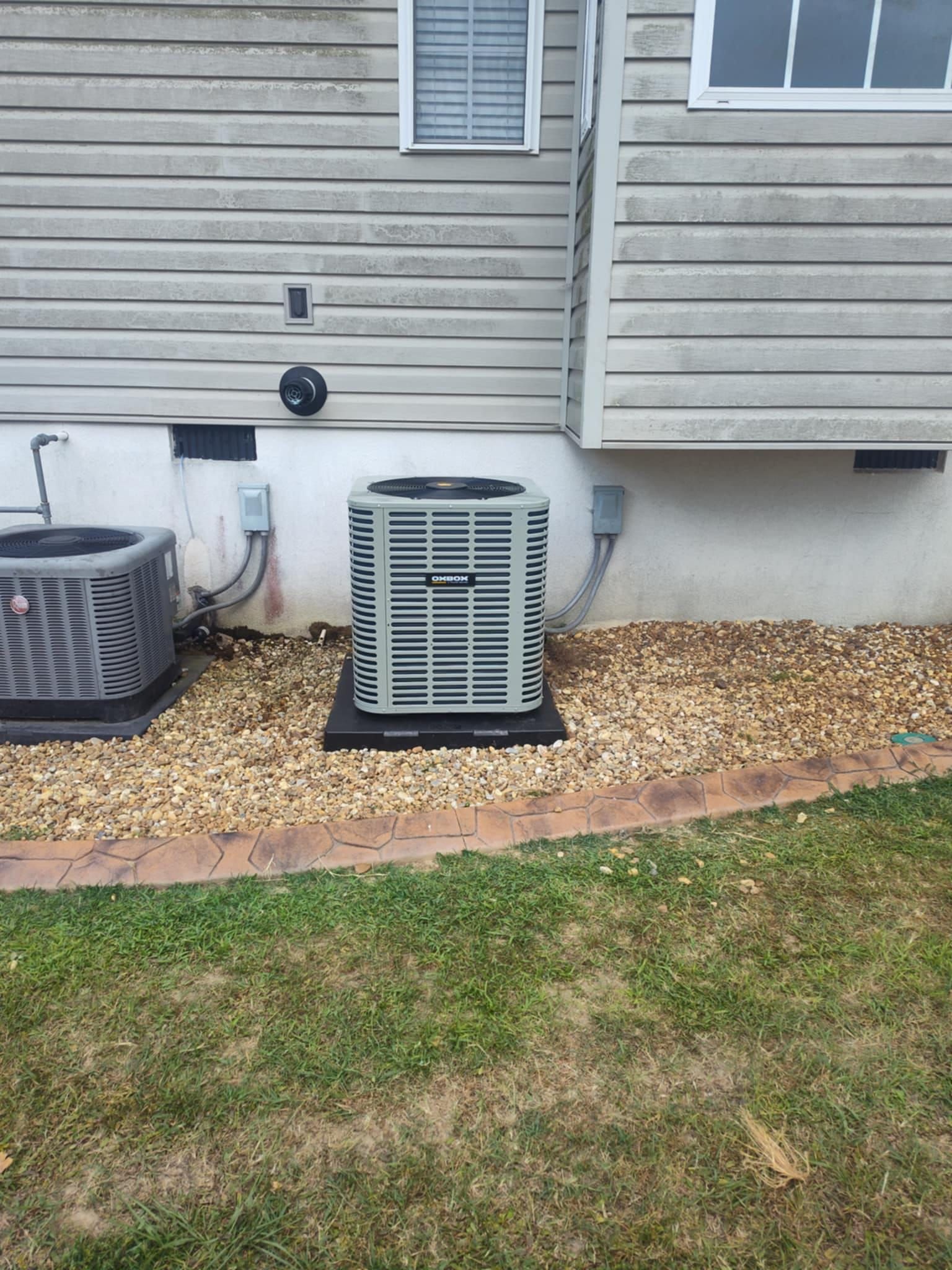 Two outdoor AC units beside a house wall, with gravel and a grass lawn in front.
