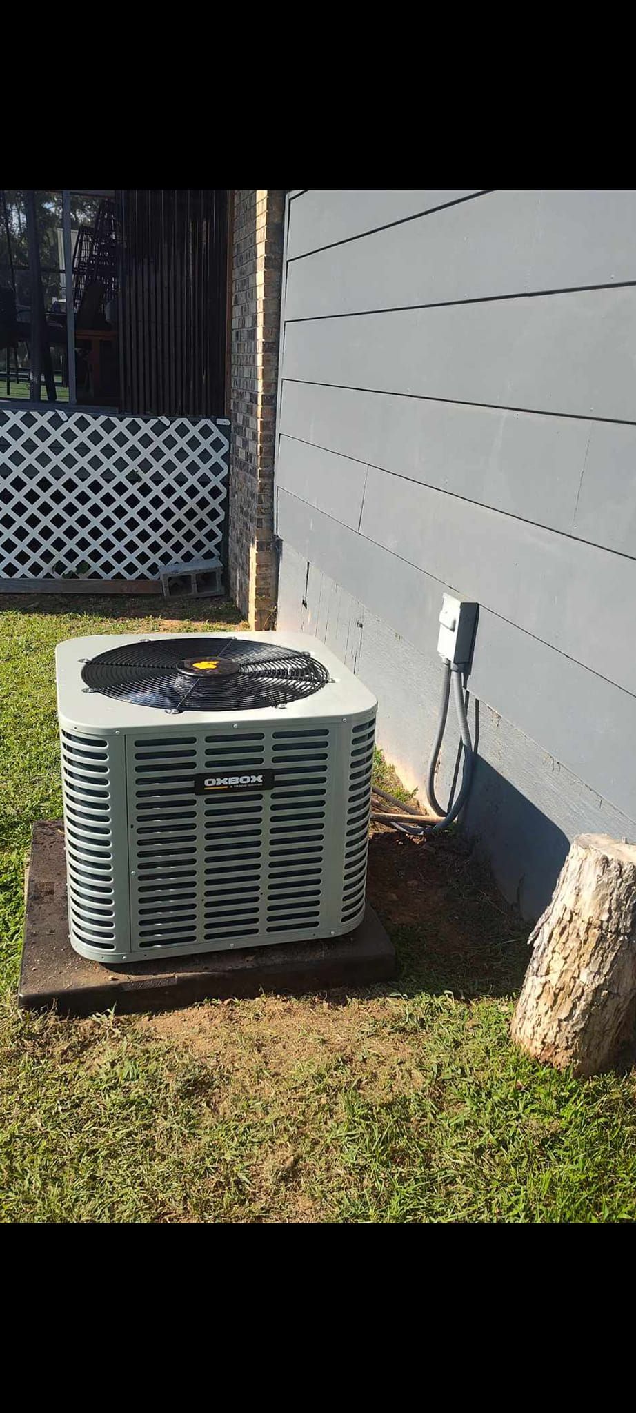 Outdoor air conditioner unit beside a house wall on a sunny day, with a lattice fence and grass nearby
