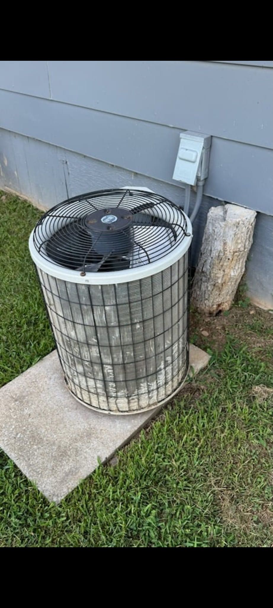 Outdoor air conditioner condenser unit beside a house wall, with a fan grill and concrete pad.