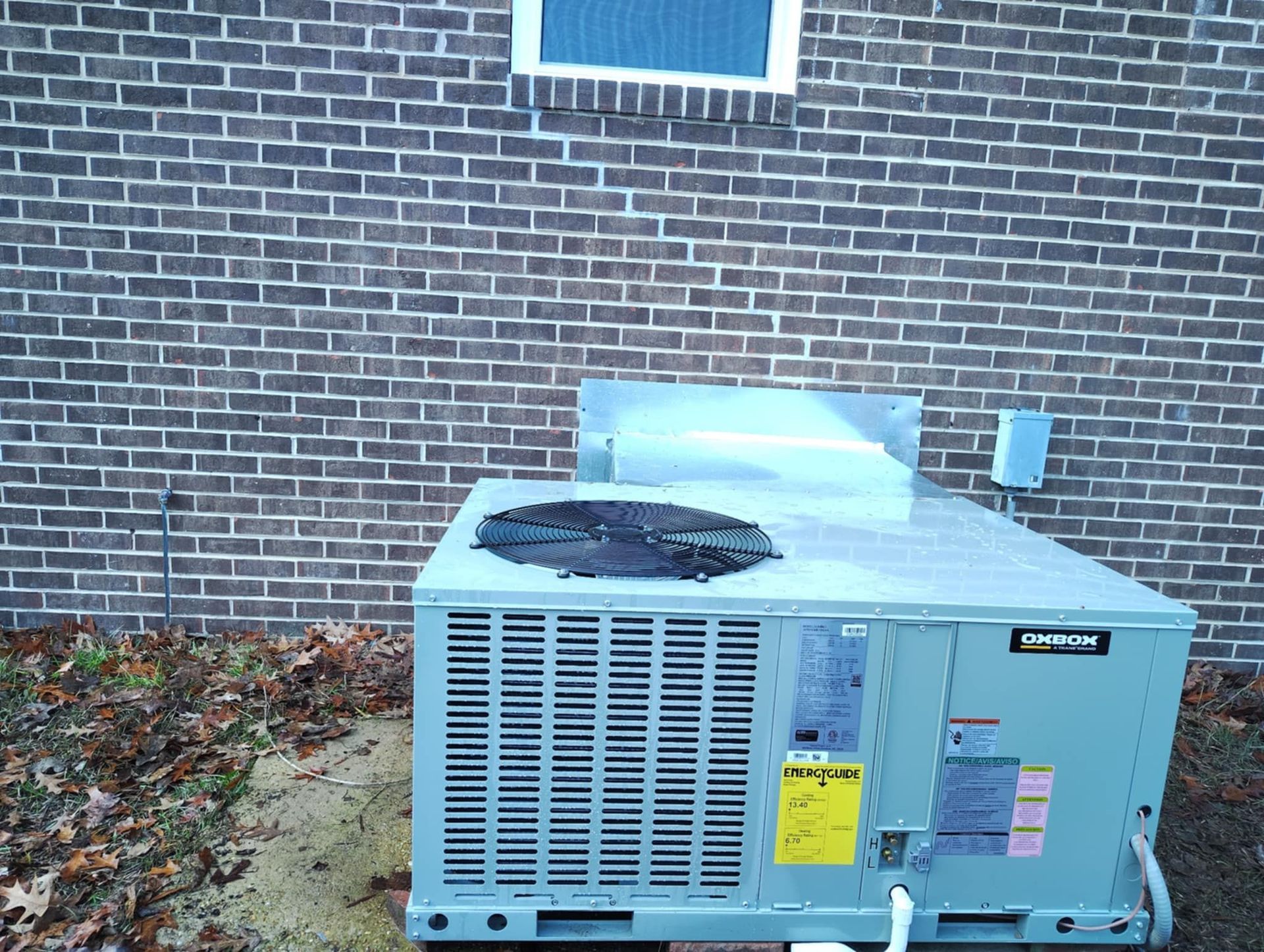 Outdoor air conditioning unit beside a brick building, with a window above and leaves on the ground