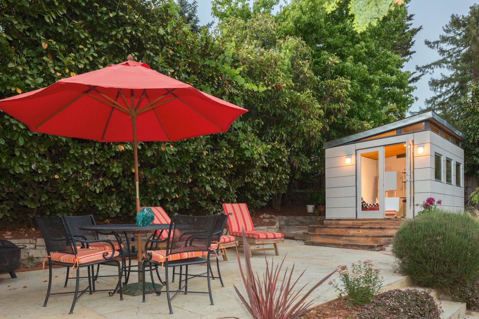 Patio with red umbrella, outdoor table and chairs, and a small lit cabin surrounded by greenery