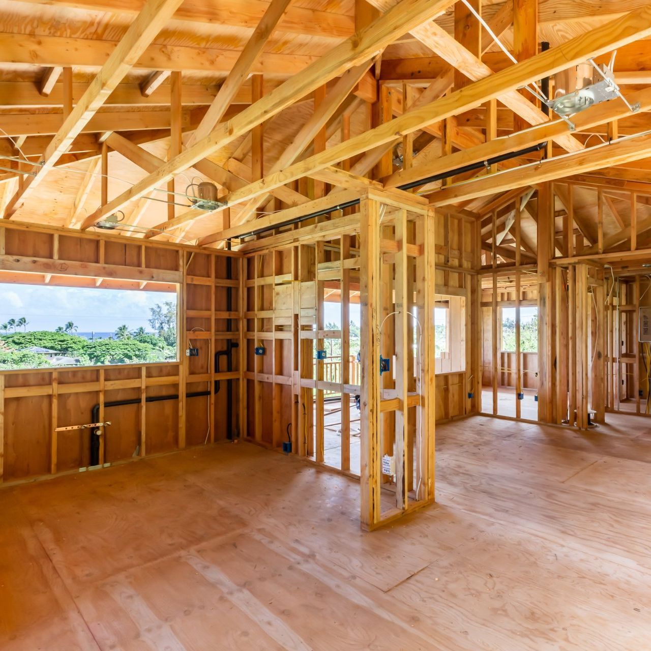 Unfinished wooden house interior with exposed framing, rafters, and open windows overlooking greenery