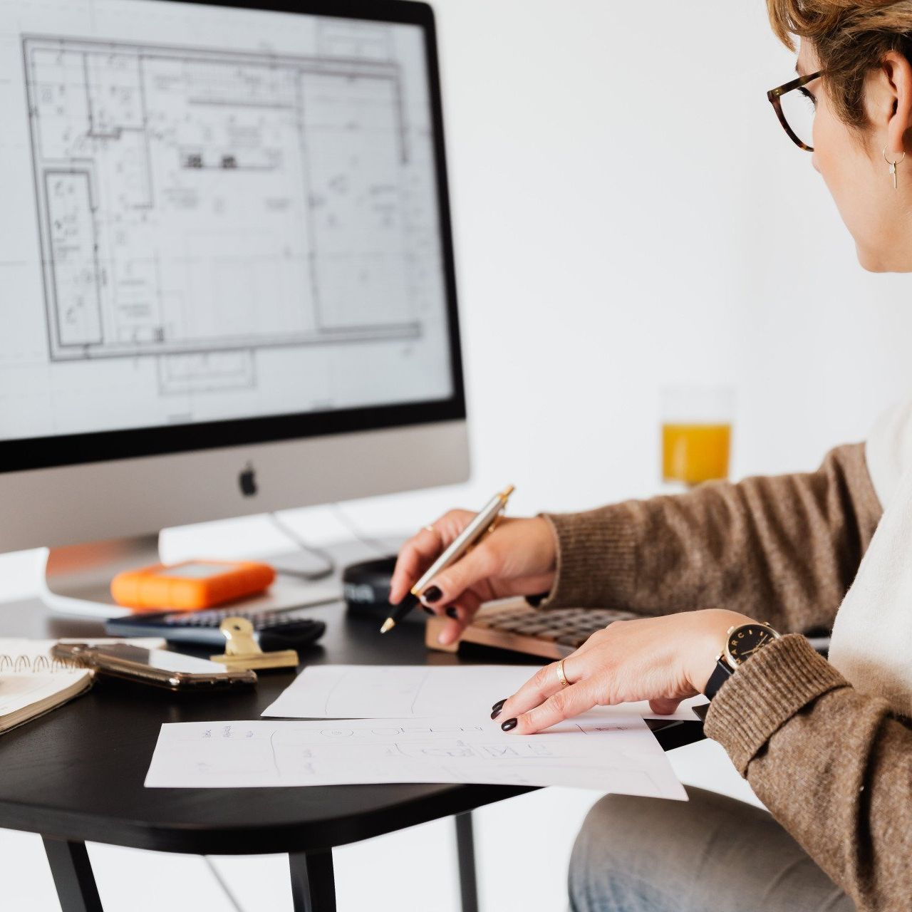 Person sketching on paper at a desk beside a computer displaying floor plans