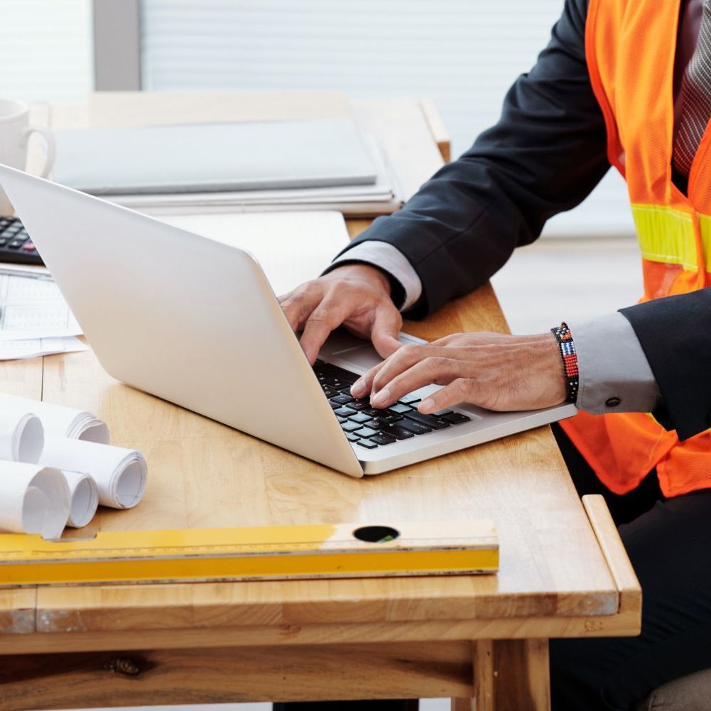 Worker in an orange safety vest typing on a laptop at a wooden desk with rolled blueprints.