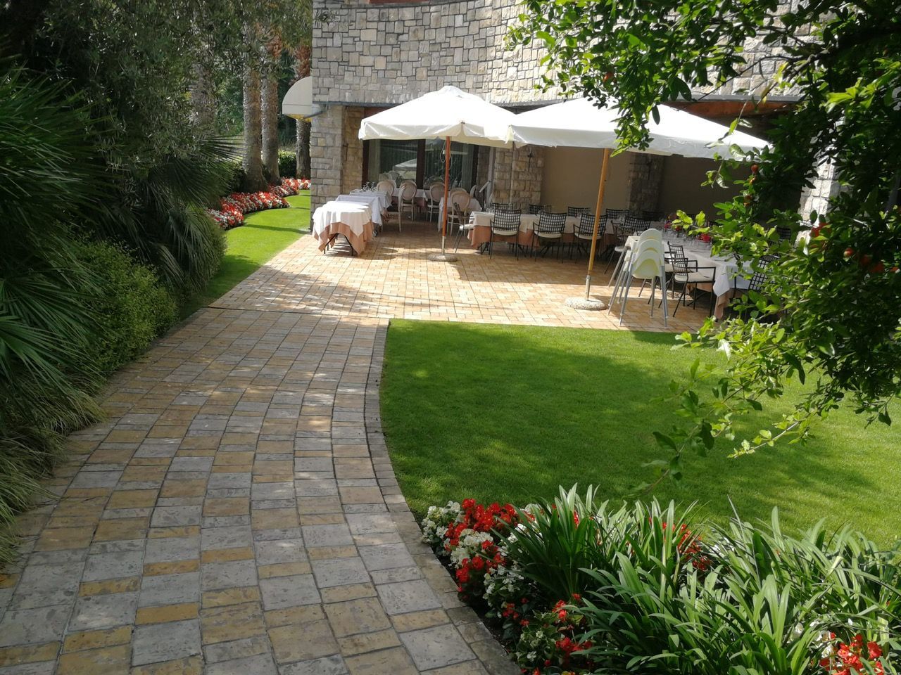 Sentiero in pietra che conduce a un patio con tavoli e ombrelloni, circondato da erba e fiori.