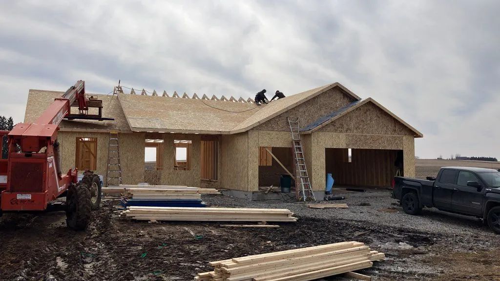 New house under construction with exposed framing, workers on roof, and trucks and lumber in front