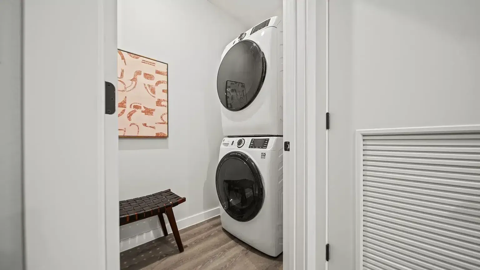 Stackable washer and dryer with a small bench and abstract art in a laundry closet.