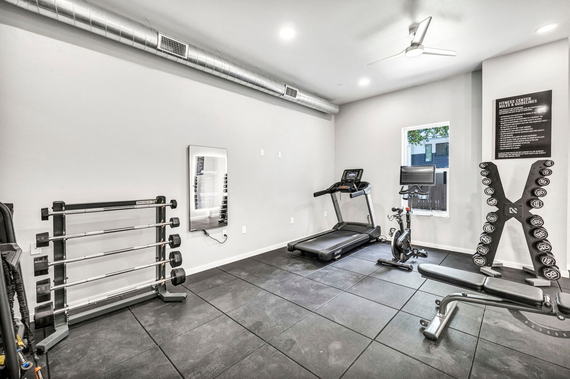 Empty home gym with treadmill, dumbbells, weight rack, and rubber flooring in a bright white room