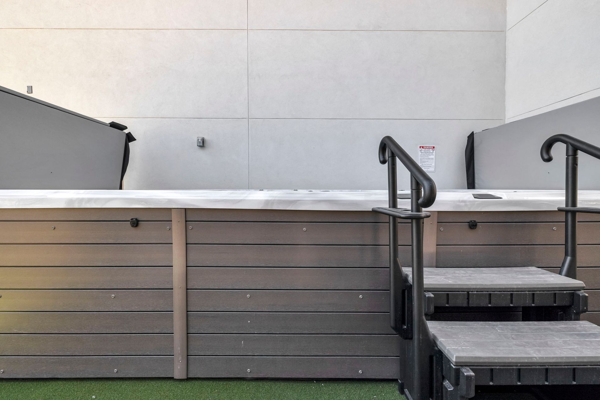 Outdoor hot tub on a deck with steps and black railings against a white wall