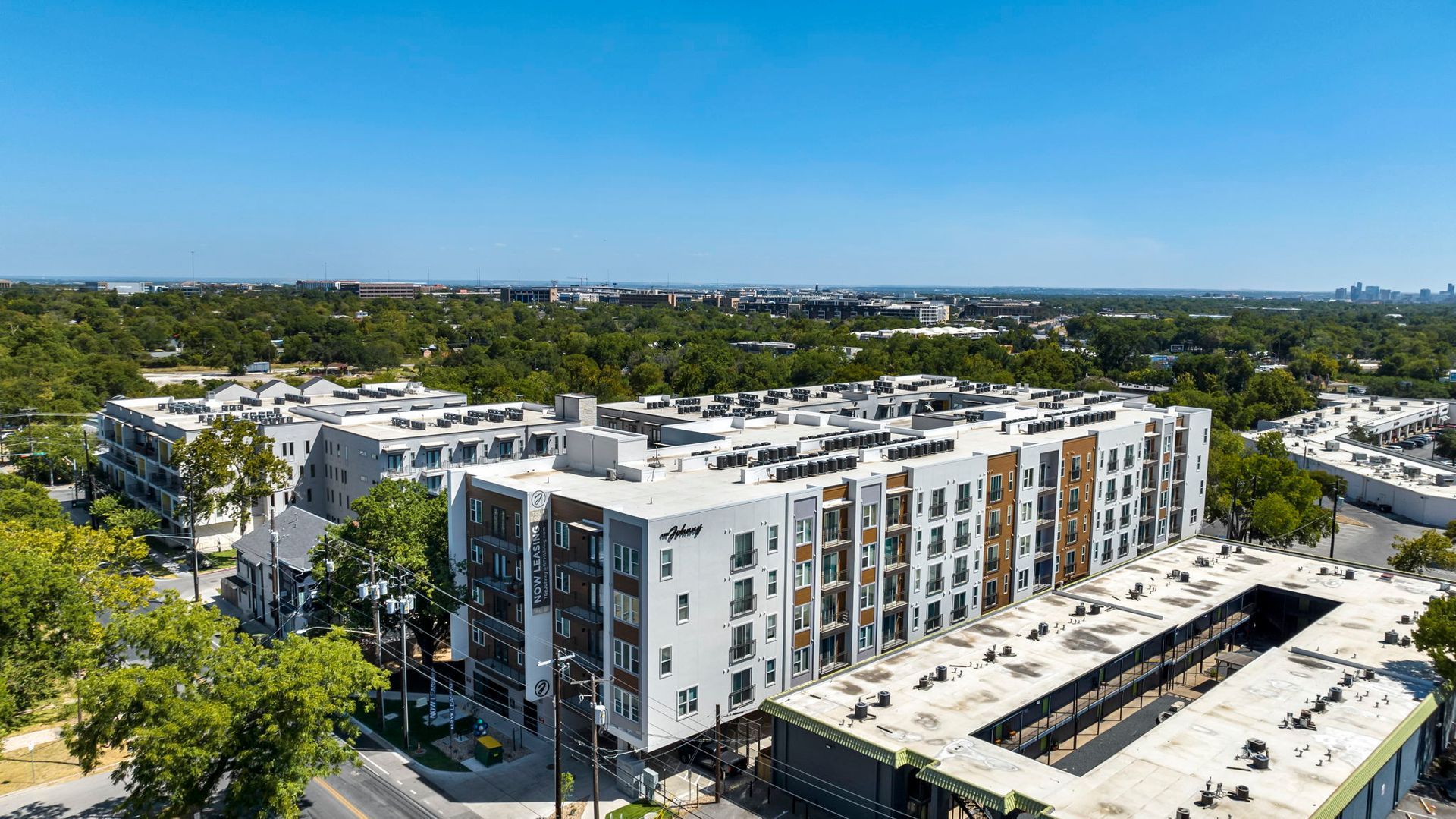 Aerial view of a modern white apartment complex surrounded by trees and nearby roads under clear skies