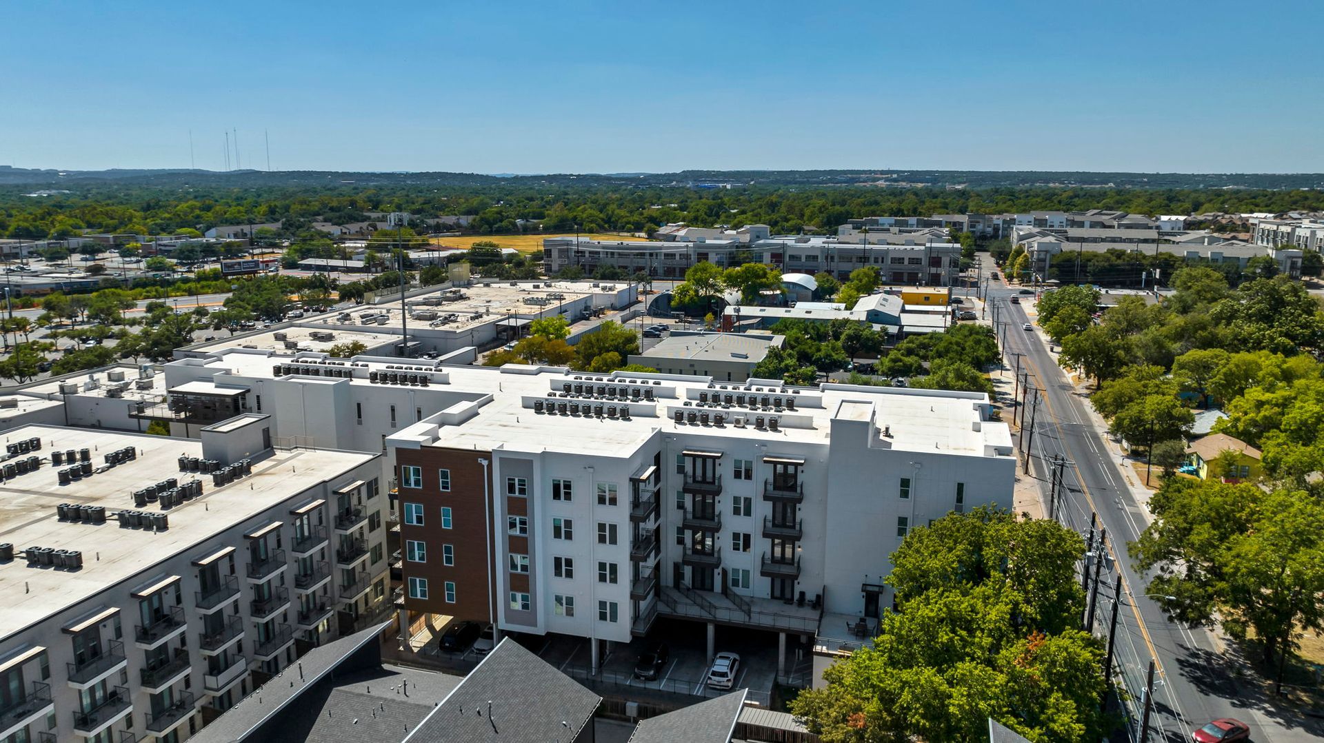 Aerial view of modern apartment buildings beside a tree-lined street under a clear blue sky