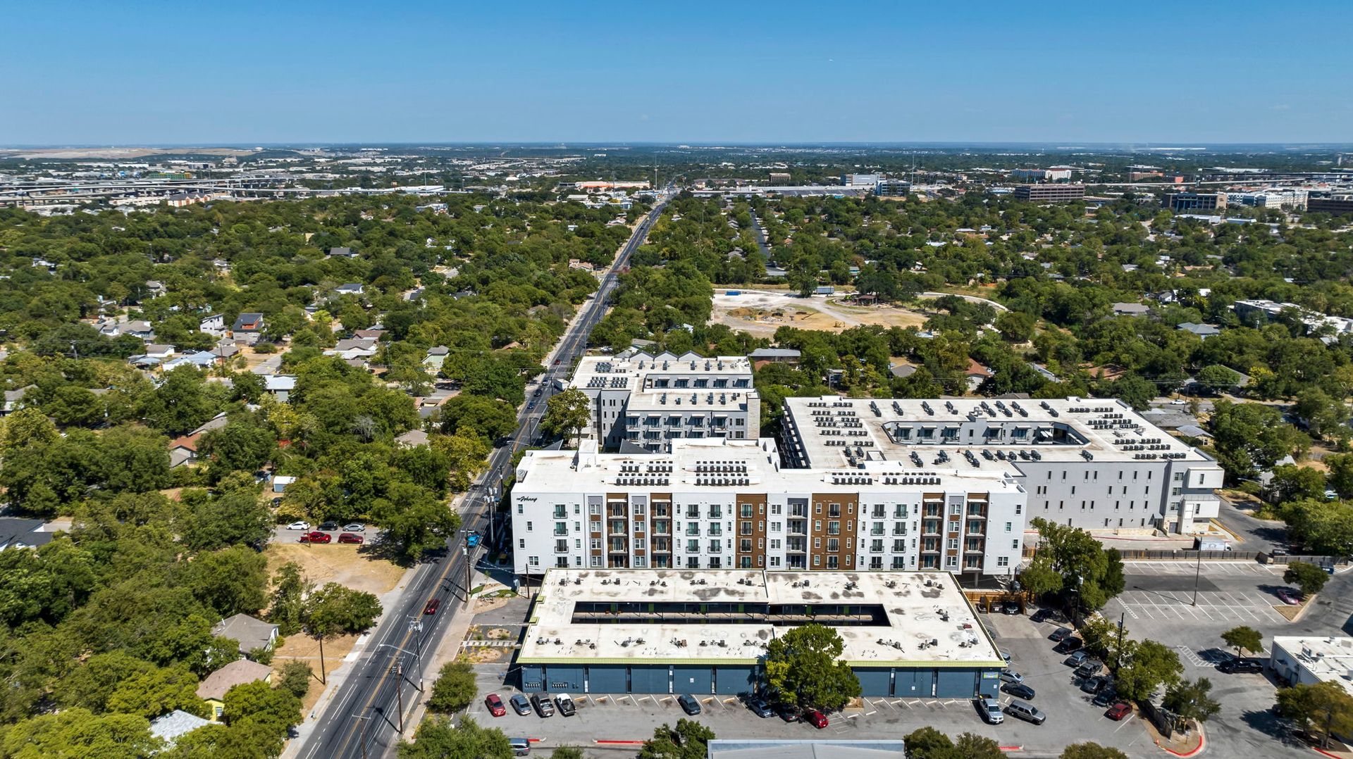 Aerial view of a white apartment complex amid green trees beside a wide road in a suburban area