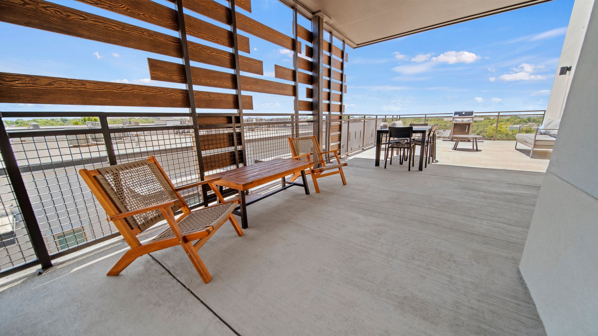 Covered rooftop patio with wooden chairs, table, and slatted shade overlooking a city view