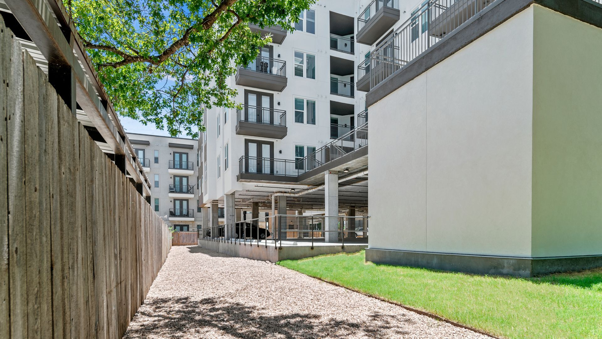 Apartment courtyard with modern buildings, gravel path, grass, and trees under a bright sky