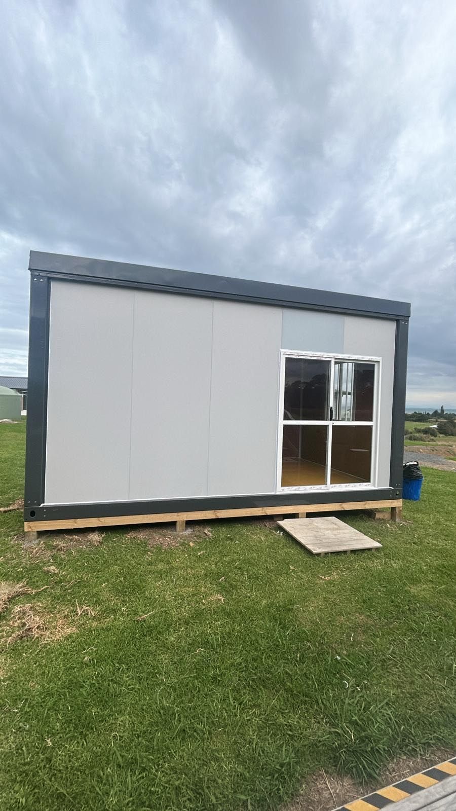 Small gray portable building on grass with a window and door under a cloudy sky.