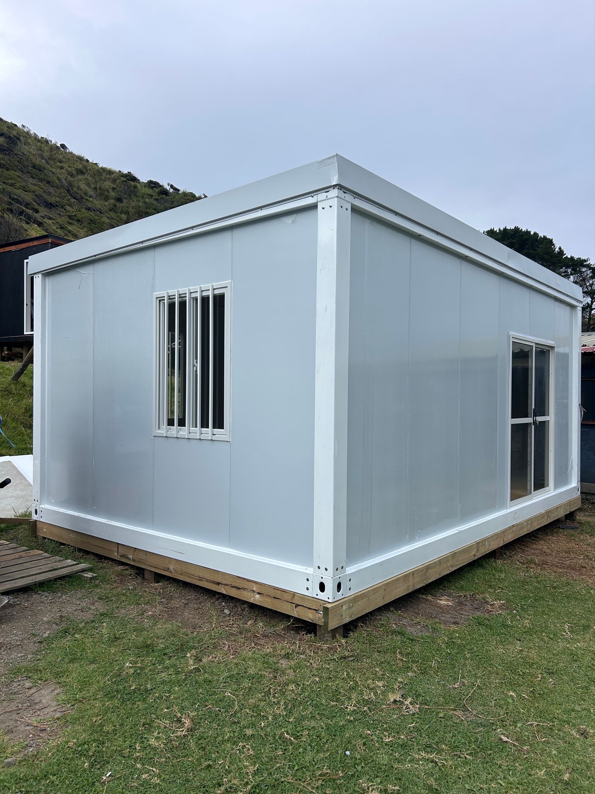 Small white modular building with windows, set on a grassy lot near a wooded hillside.