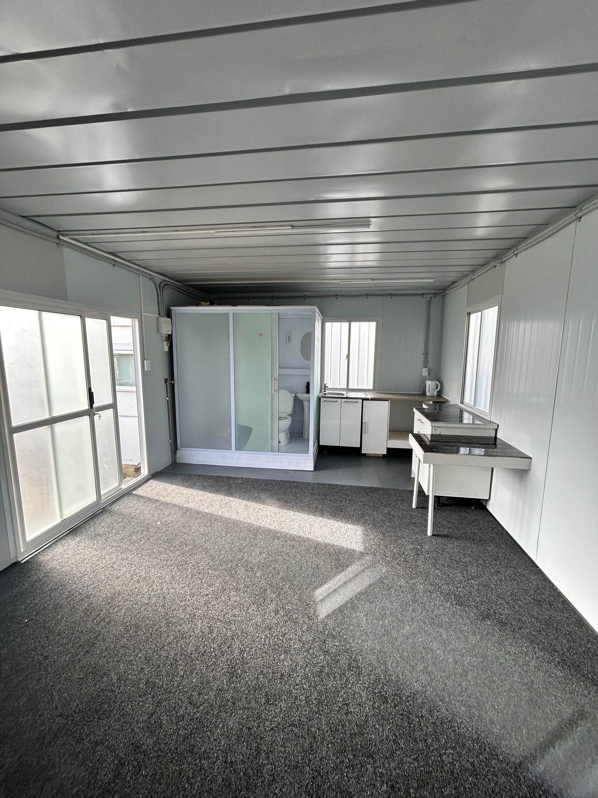 Empty narrow room with white walls, gray speckled floor, windows, and a desk along the right side.