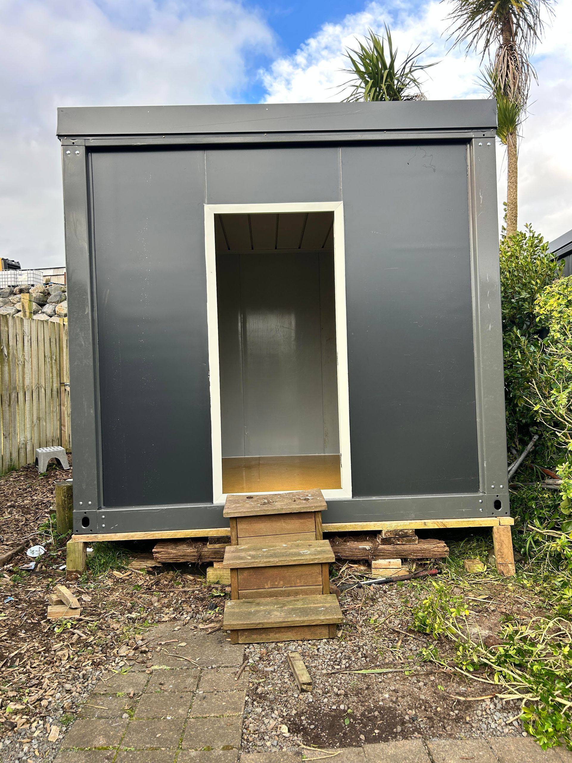 Small gray shed with open doorway and wooden steps in a backyard, surrounded by grass and gravel.