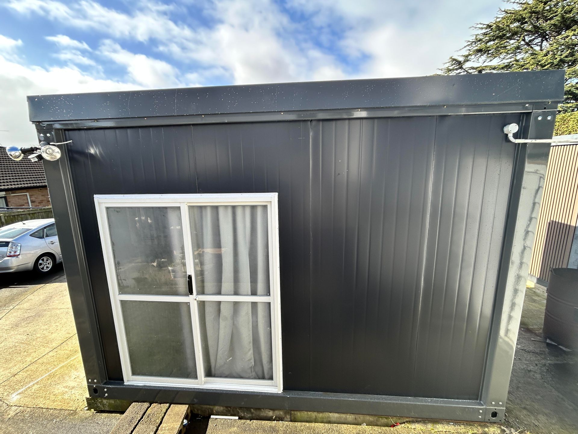 Small dark gray modular shed with a white-framed window and door-like panel on a paved lot