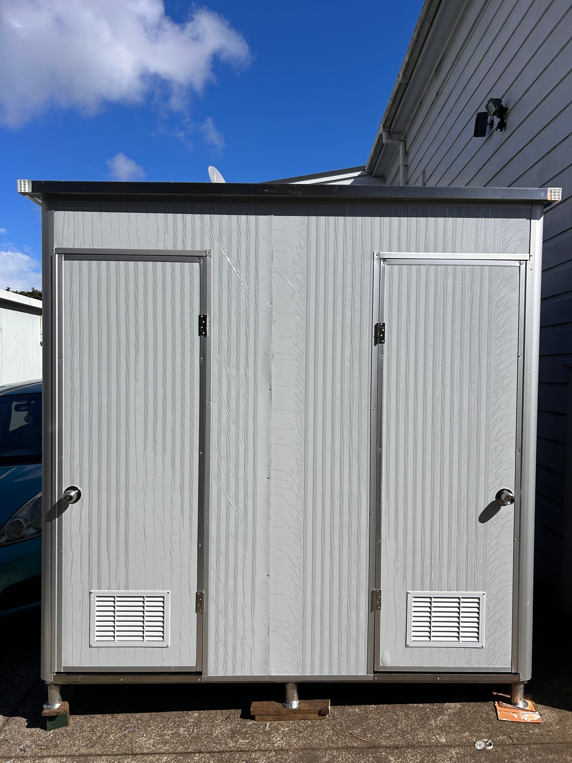White utility shed with two doors and vents, standing outdoors beside a larger building under a blue sky.