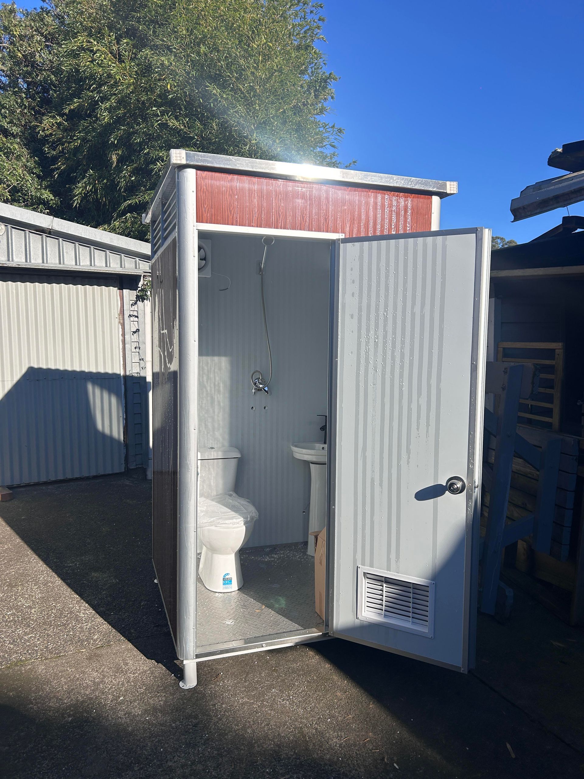 Outdoor portable toilet stall with open white door, showing a toilet inside beside a fence and gravel yard.