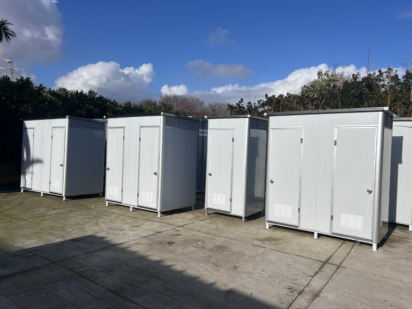 Row of white portable toilets on a paved lot under a blue sky with clouds