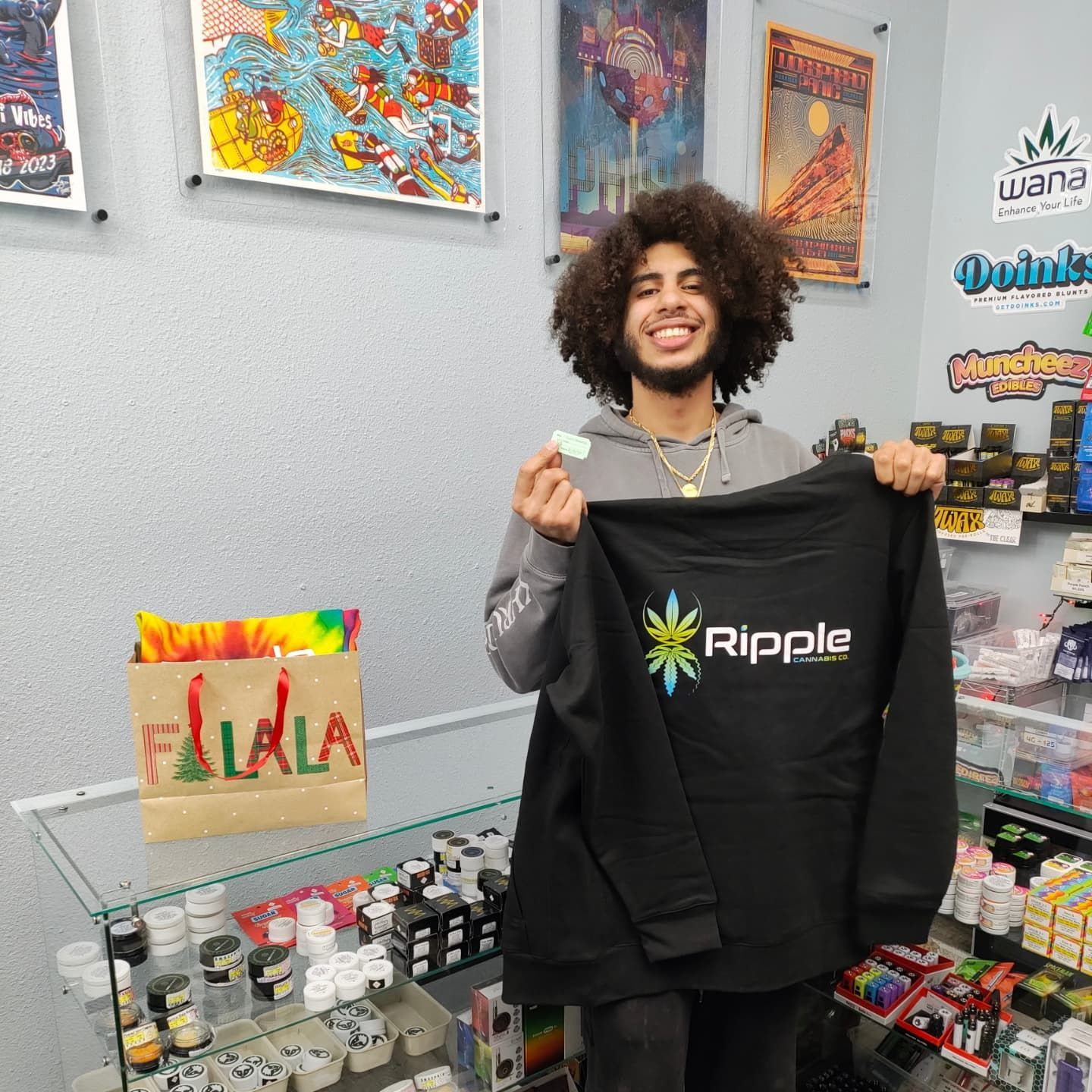 Person holding Ripple hoodie and treat in a shop, smiling. Behind them are products and art.