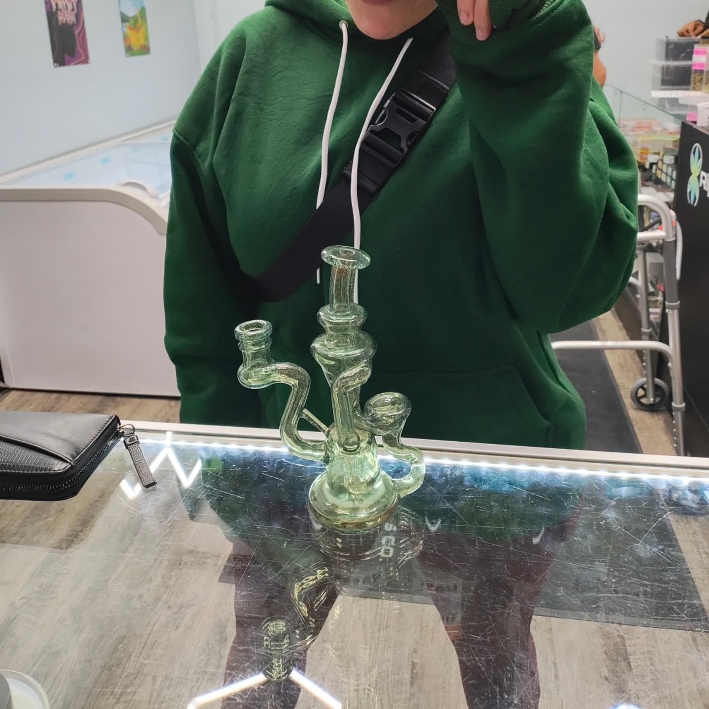 Person in green hoodie with glass bong on a glass display case inside a store.