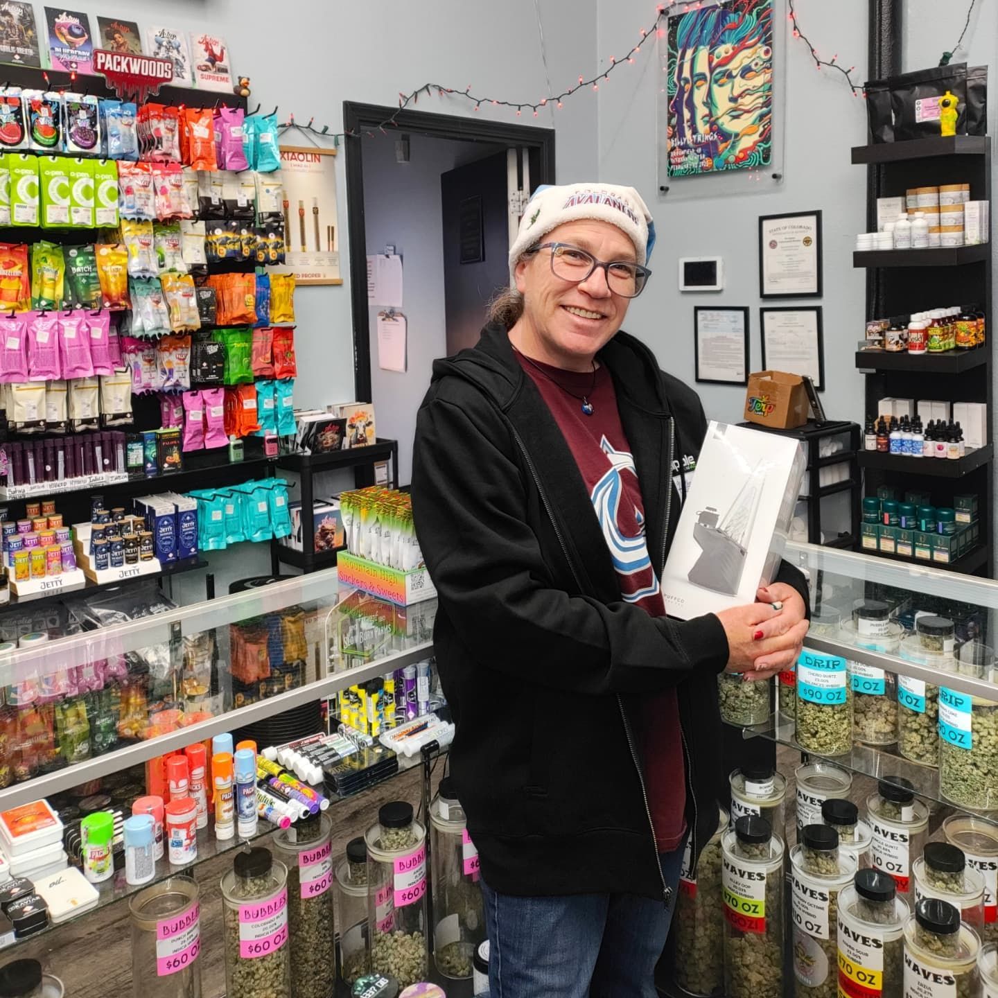 Person holding a bag smiles in a cannabis store. Displays of products, jars of green, and colorful packaging visible.