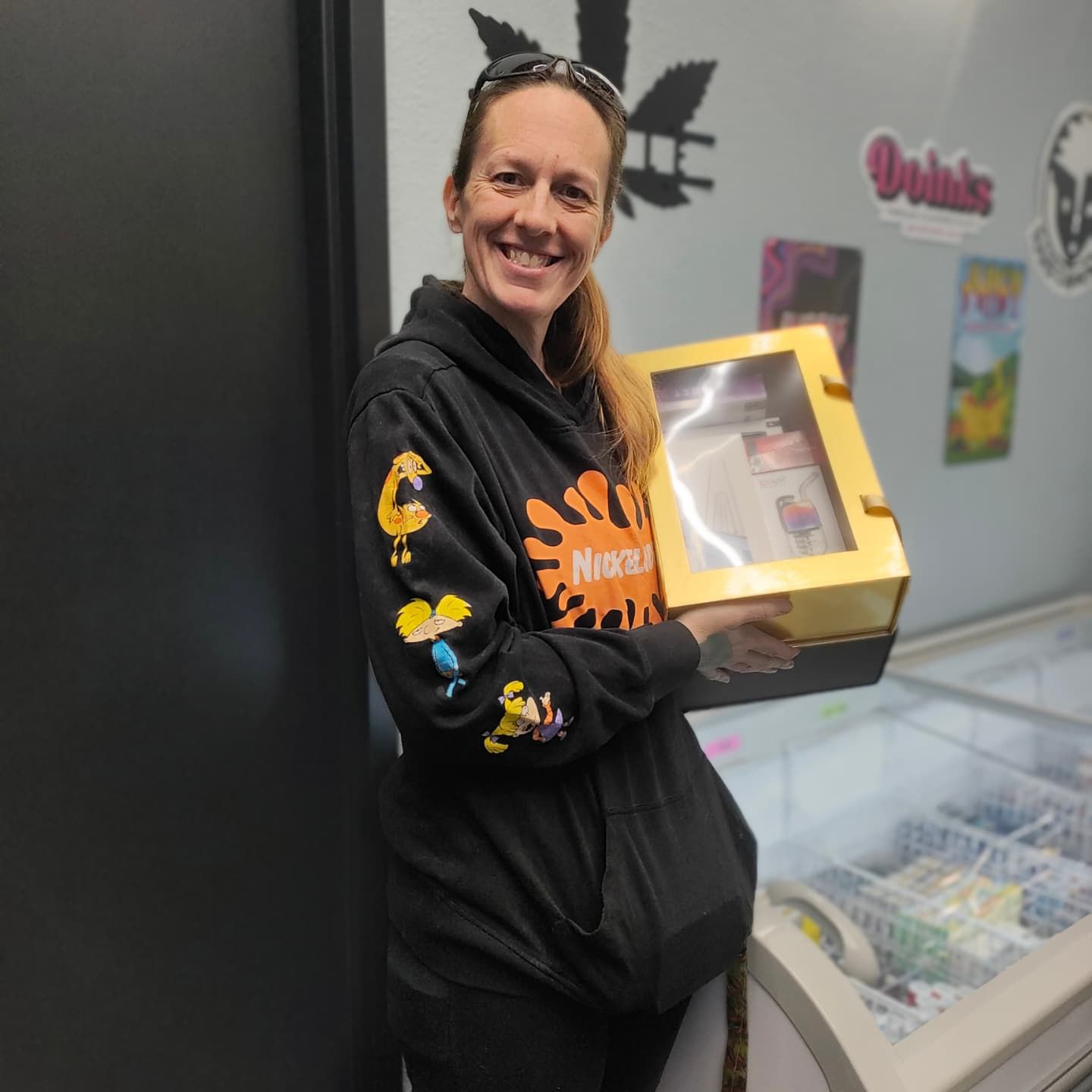 Woman in black hoodie holding a gold gift box, smiling, in front of a display case.