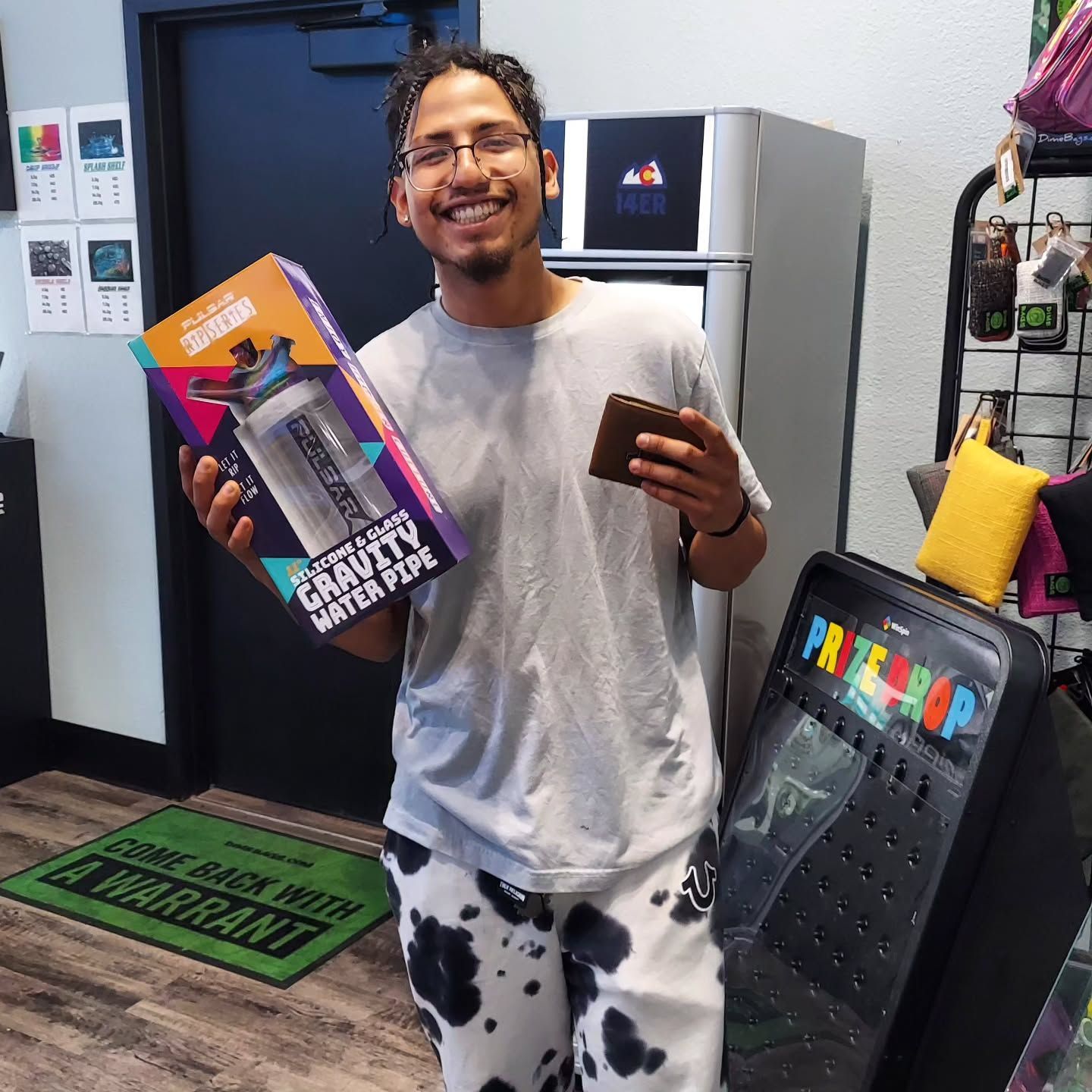 Man holding a water pipe and wallet, smiling. Inside a shop with a prize machine.