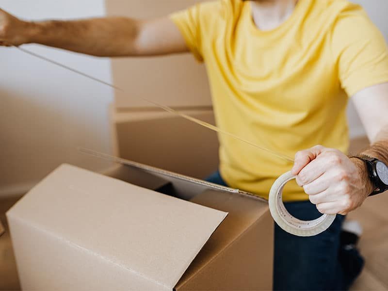 A man in a yellow shirt is packing a cardboard box with tape.