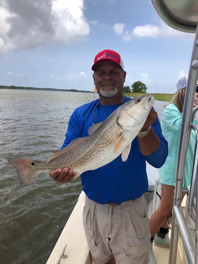 Man holding a large fish on a boat, wearing a blue shirt and red cap on a cloudy lake.