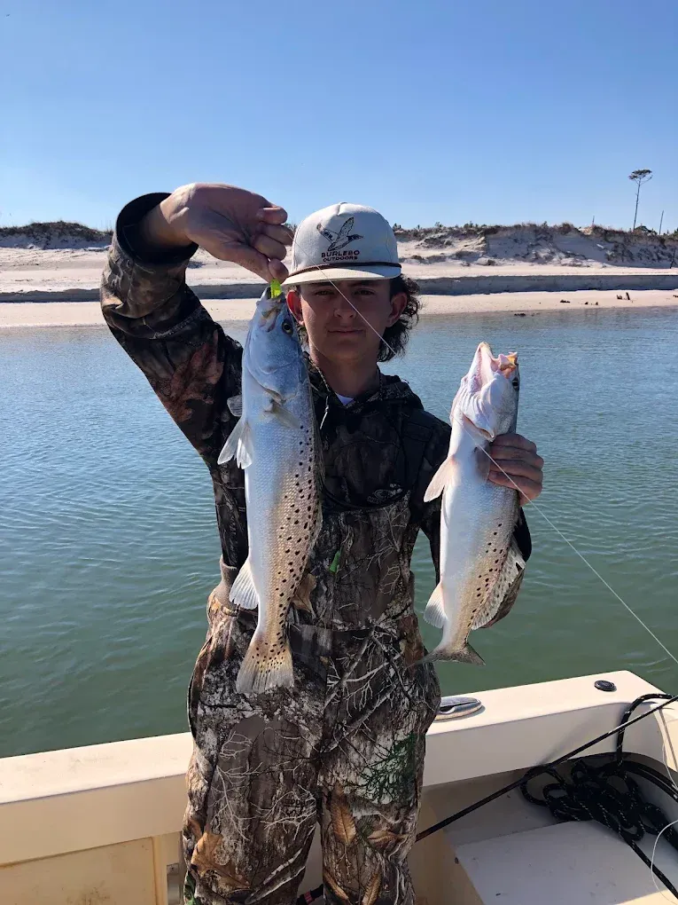 Angler on a boat holding two large fish, with a sandy shoreline and water in the background.