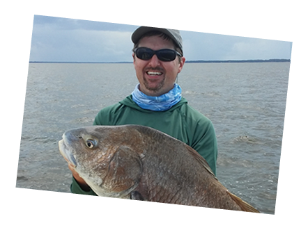 Man holding a large fish in a boat on calm water, wearing sunglasses and a cap.