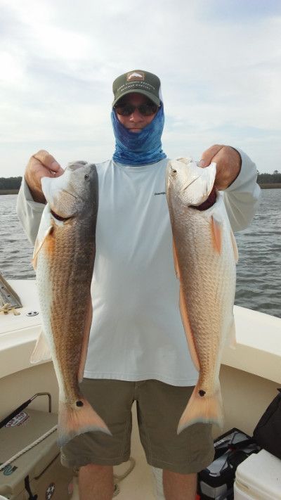 Person on a boat holding two large fish over the water, wearing a cap and face covering.