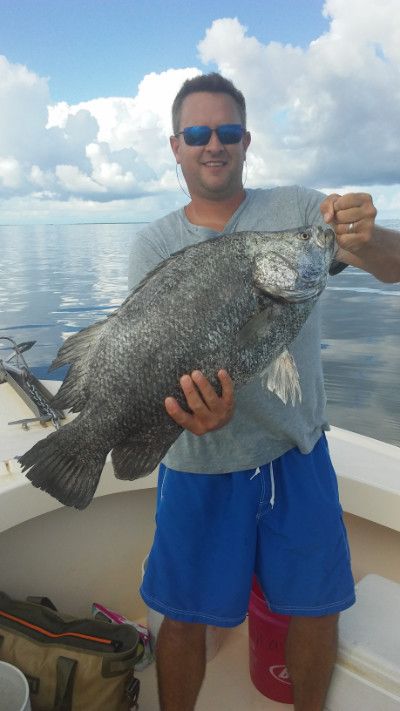Man holding a large fish on a boat under a cloudy sky, wearing sunglasses and blue shorts.