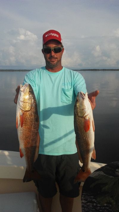 Man holding two large fish on a boat, wearing a red cap and teal shirt with calm water behind him