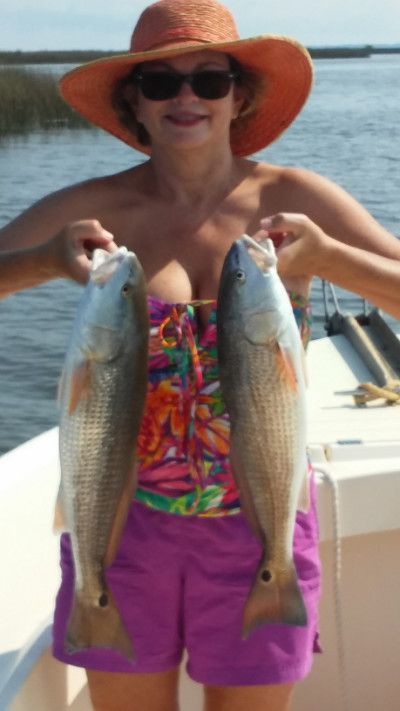 Woman on a boat holding two large fish, wearing a sunhat, sunglasses, colorful top, and purple shorts.