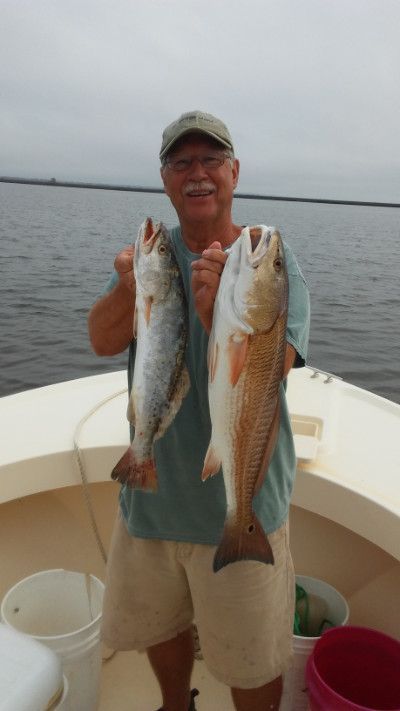 Man on a boat holding two large fish, wearing a cap and smiling on calm water under a cloudy sky