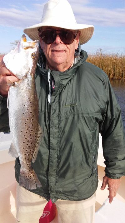 Man holding a large fish on a boat, wearing a white hat, sunglasses, and green jacket.