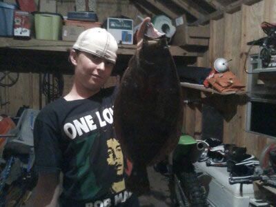 Person in a hard hat holds up a large dark fish in a cluttered garage or workshop.