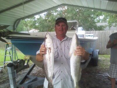 Man holding two large fish under a covered outdoor area, with a boat in the background