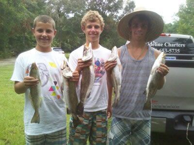 Three people holding up fish outdoors near a parked truck, smiling after a fishing trip.