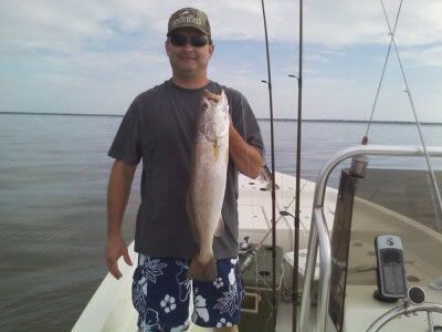 Man holding a large fish on a boat deck with fishing rods and calm water in the background