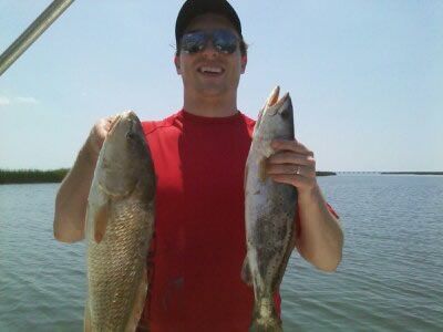 Man holding two large fish at a lakeside, wearing a red shirt and cap.
