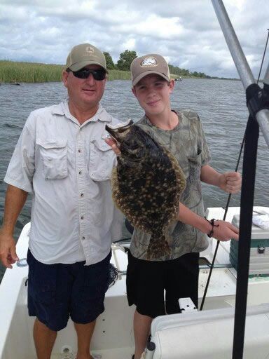 Two people on a boat holding a large fish, with water and marshland in the background.