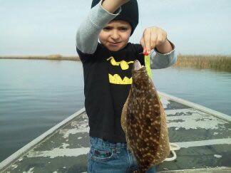 Child holding up a fish on a dock beside calm water, wearing a black shirt and gray hat.