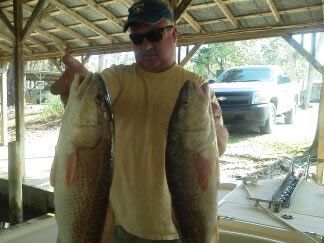 Man holding two large fish under a carport, with a truck and boat in the background.