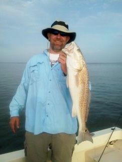 Man holding a large fish on a boat, wearing a light blue shirt and hat on calm water.